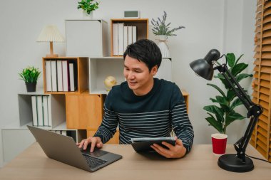 Ev ofisinde dizüstü bilgisayar ve dijital tabletle çalışan mutlu adam.