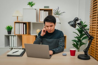 Genç adam ev ofisinde dizüstü bilgisayar ve kablosuz kulaklıkla çalışıyor.