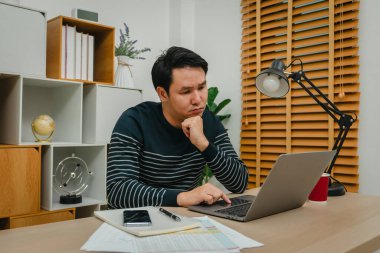 Genç adam evdeki bilgisayarla düşünüyor ve çalışıyor.