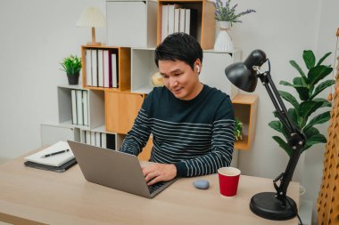 Genç adam ev ofisinde dizüstü bilgisayar ve kablosuz kulaklıkla çalışıyor.