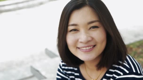 Gros plan portrait de jeune femme asiatique souriant avec des cheveux noirs soufflant dans le vent regardant et flou fond ralenti — Video