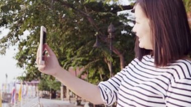 Genç Asyalı gülümseyen ve günbatımı Nehri yakınında bakarak rüzgarda üfleme siyah saçlı bir smartphone kullanarak kadın portresi kapatın. Giyen kadın ekose gömlek siyah ve beyaz olduğunu. Ağır çekim.