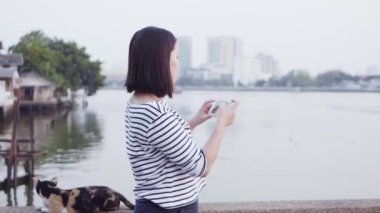 Genç Asyalı gülümseyen ve günbatımı Nehri yakınında bakarak rüzgarda üfleme siyah saçlı bir smartphone kullanarak kadın portresi kapatın. Giyen kadın ekose gömlek siyah ve beyaz olduğunu. Ağır çekim.