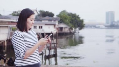 Genç Asyalı gülümseyen ve günbatımı Nehri yakınında bakarak rüzgarda üfleme siyah saçlı bir smartphone kullanarak kadın portresi kapatın. Giyen kadın ekose gömlek siyah ve beyaz olduğunu. Ağır çekim.