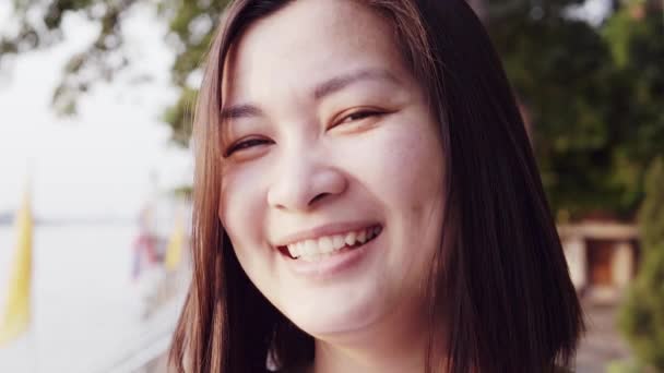 Close up retrato da jovem mulher asiática sorrindo com o cabelo preto soprando no vento olhando para o pôr do sol perto do rio. Mulher vestindo é xadrez camisa preto e branco com cena Slow Motion . — Vídeo de Stock