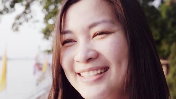 Close up retrato da jovem mulher asiática sorrindo com o cabelo preto soprando no vento olhando para o pôr do sol perto do rio. Mulher vestindo é xadrez camisa preto e branco com cena Slow Motion . — Vídeo de Stock