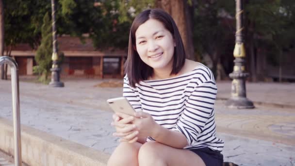 Närbild porträtt av ung asiatisk kvinna leende och med en smartphone med svart hår som blåser i vinden och tittar på solnedgången nära floden. Kvinna som bär är rutig skjorta svart och vitt. Slow Motion. — Stockvideo