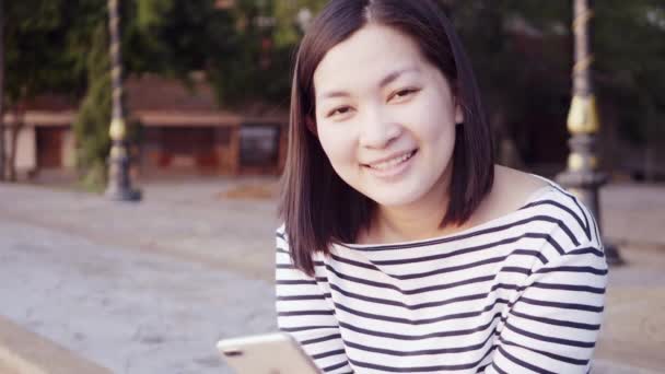 Close up portrait of Young Asian Woman smiling and using a smartphone with Black hair blowing in wind looking at the sunset near the river. Woman wearing is plaid shirt black and white. Slow Motion. — Stock Video