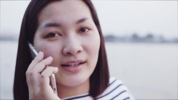 Bliska portret kobiety Azjatki młode uśmiechając się i za pomocą smartfona z czarne włosy wiejący wiatr, patrząc na zachód, w pobliżu rzeki. Kobieta ubrana jest plaid shirt czerni i bieli. Zwolnionym tempie. — Wideo stockowe