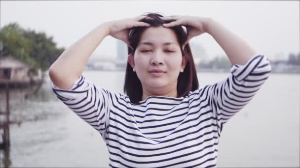 Zavřete portrét mladé asijské ženy s úsměvem a pomocí rukou masírovat hlavu s černými vlasy vlající ve větru, při pohledu na západ slunce v blízkosti řeky. Žena nosí je kostkované košile. Zpomalený pohyb. — Stock video