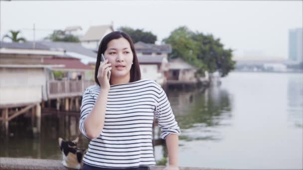 特写的年轻亚洲妇女微笑和使用智能手机与黑头发吹在风中看着河边的日落。穿的是格子衬衫的黑白。慢动作. — 图库视频影像