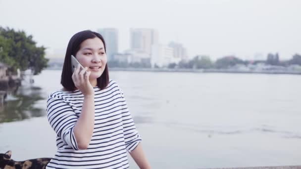 特写的年轻亚洲妇女微笑和使用智能手机与黑头发吹在风中看着河边的日落。穿的是格子衬衫的黑白。慢动作. — 图库视频影像