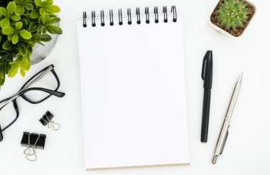 Boş defter sayfası ve malzemeleri olan beyaz ofis masası. Üst manzara, düz uzanma.