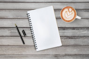 Boş defter ahşap masanın üstünde, içinde kalem ve kahve var. Üst manzara, düz uzanma.
