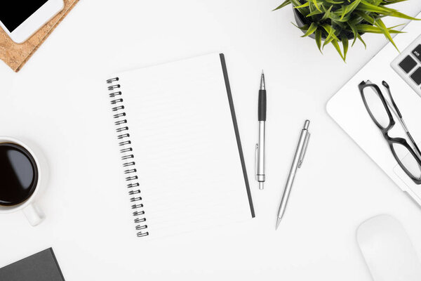 Чистый блокнот лежит на столе белого офиса с ноутбуком, кофейной чашкой и офисными принадлежностями. Вид сверху с копировальным пространством, плоская кладка.