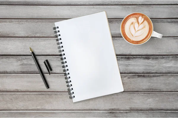 Boş defter ahşap masanın üstünde, içinde kalem ve kahve var. Üst manzara, düz uzanma.