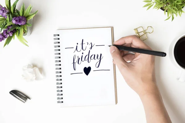 Man is writing it's friday calligraphy text on the notebook on white office desk table. Top view, flat lay.