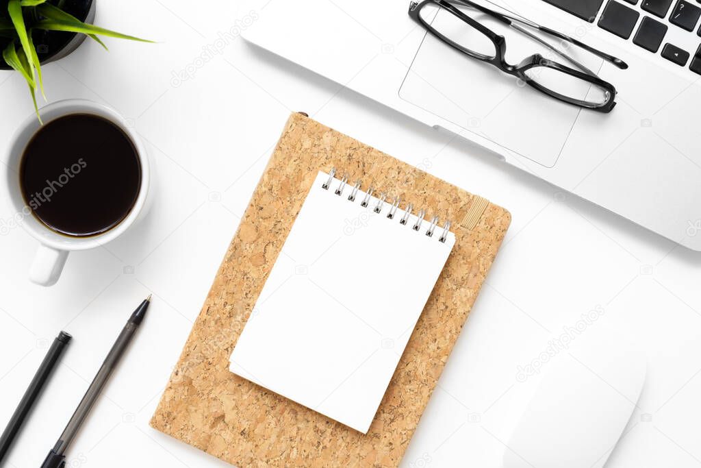 White office desk table with blank notebook, laptop, cup of coffee and office supplies. Top view with copy space, flat lay.