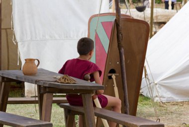 Aquileia, İtalya - 22 Haziran 2014: çadır ve yerel yıllık antik Roma tarihi canlandırma kalkanlar arasında ahşap bir bankta oturmuş bir kostümlü genç çocuk