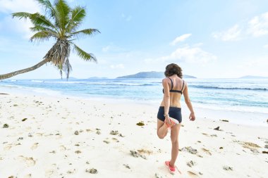 Genç bir kadın tropikal plajda egzersiz yapmadan önce geriniyor.