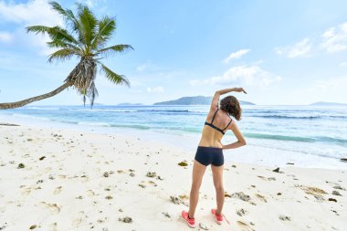 Genç bir kadın tropikal plajda egzersiz yapmadan önce geriniyor.