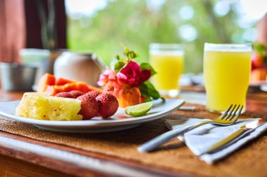 Bir bardak meyve suyuyla eşleştirilmiş taze tropikal meyveler, ananas, papaya ve karpuz içeren renkli bir kahvaltı. Sağlıklı beslenme, egzotik tatlar ve sabah rahatlama temaları için idealdir.