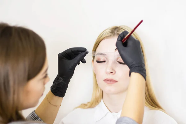 Female cosmetologist performs eyebrow correction on beautiful models in the beauty parlor. The girl's face is a blonde. Close-up. Facial care — Stock Photo, Image