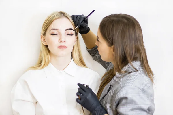 美容師は美容院で美しいモデルに眉の補正を行います。その少女はブロンドだ。顔面治療ヘナ染色 — ストック写真