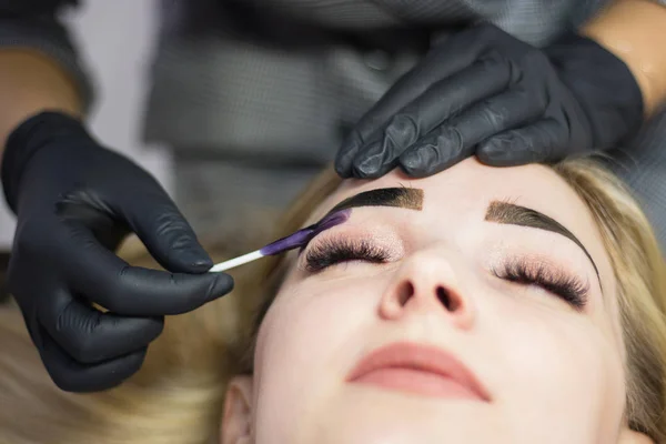 Beautician performs eyebrow correction on a beautiful model in a beauty salon. The girl is blonde. Facial treatment. Henna dyeing