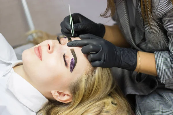 Beautician performs eyebrow correction on a beautiful model in a beauty salon. The girl is blonde. Facial treatment. Henna dyeing