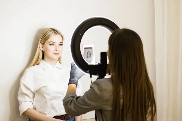 O esteticista fotografa o seu trabalho num telemóvel. Duas raparigas num salão de beleza. Lâmpada de anel para maquiadores. Cuidado com o rosto — Fotografia de Stock