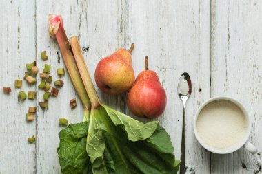 Rubarb sapları ve beyaz ahşap bir arka planda iki armut. Yakınlarda bir kupa şeker ve metal kaşık vardı. Tamamlayıcı renkler