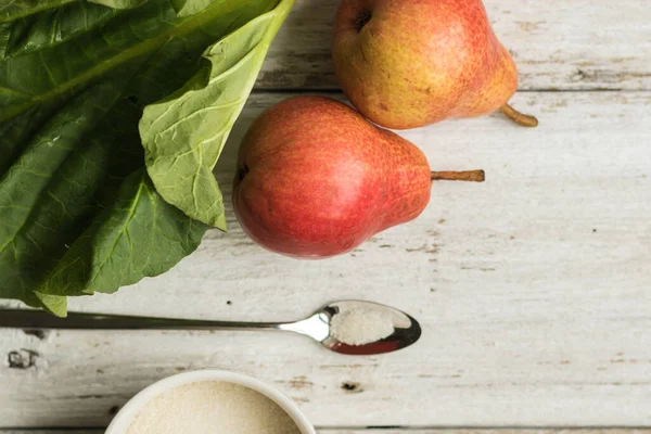 Steli di rabarbaro e due pere su uno sfondo di legno bianco. Un boccale di zucchero e un cucchiaio di metallo si trovano nelle vicinanze. Colori complementari — Foto Stock