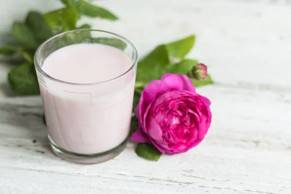 Аюрведический напиток розовое молоко или маття. Стакан освежающего коктейля стоит на деревянном белом фоне. Рядом находится лиловая роза. — стоковое фото