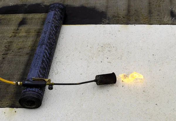 Material and equipment for waterproofing roofs and terraces. Roll of bituminous sheath, propane torch and glove resting on a bituminous membrane already laid.