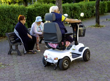 Modugno, İtalya - 11 Mayıs 2018: Yaşlılar bir park bankında sosyalleşirler. Engelli bir kişi motorlu tekerlekli sandalye kullanır