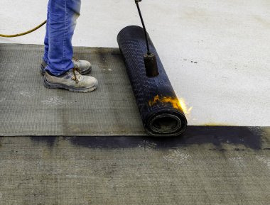 Bitumen çatı keçesinin bir kısmını ısıtıcı meşale ateşiyle eritmek için hazırlayan işçiler. 