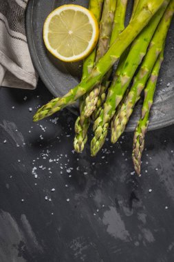 Bohça-in genç çiğ çiğ organik yeşil kuşkonmaz dilimlenmiş limon ve siyah doku arka plan üzerinde deniz tuzu. Üstten Görünüm. Sağlıklı beslenme