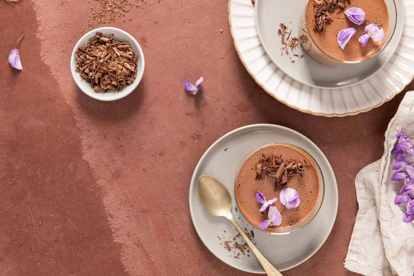 Tepeden bakıldığında, kaşık, talaş ve kırsal bir yüzeyde mor çiçeklerle şekillendirilmiş iki lezzetli çikolata mus görülüyor..