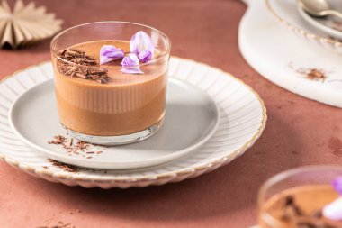 Çikolata parçacıkları ve mor çiçeklerle süslenmiş bir bardak kremalı çikolatalı mus, bir tabakta şekillendirilmiş, cezbedici bir tatlı.