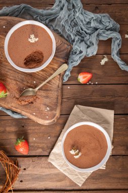 Zarif beyaz çiçeklerle süslenmiş iki kase yumuşak çikolatalı mus gümüş kaşıkla kırsal ahşap bir masada sunuluyor.