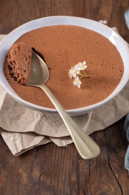 Beyaz bir kasede kadife çikolatalı mus, porsiyonunu kepçeleyen altın bir kaşık, narin beyaz çiçeklerle süslenmiş, kırsal ahşap bir masanın üzerinde buruşuk parşömen kağıdına konmuş.