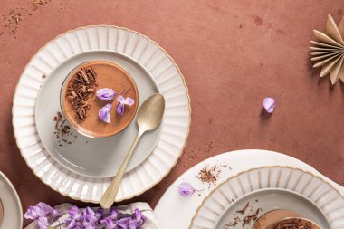 Gözlüklü iki kremalı çikolatalı mus, bir kaşık, çiçekler ve kırsal bir yüzey üzerindeki dekoratif elementlerden oluşan genel bir manzara..