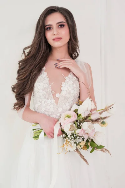 Slim beautiful woman wearing luxurious wedding dress near the panoramic window. Gorgeous bride holding flowers. — Stock Photo, Image