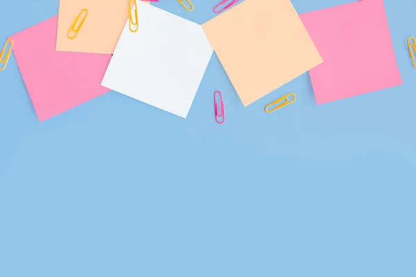 Square colored sticky notes and paper clips on a blue background. Flatlay with copy space
