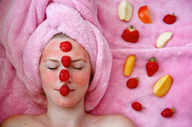 Pembe havlusu olan bir kız yüzünde çilek maskesiyle gözleri kapalı yatar. Meyve parçalarının yanında