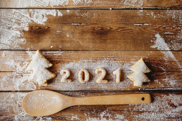 Bakery Flat lay with cookies in form of Christmas trees and numbers 2021, flour,wooden spoon on the wooden table. Copyspace.