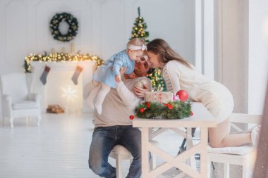 Masadaki beyaz odada bir aile. Yeni yıl havası. Arka planda süslü şömine ve Noel ağacı var.