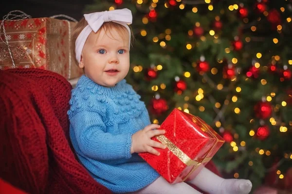 Una bambina con un vestito blu a maglia si siede e tiene una confezione regalo rossa. Bokeh sfondo dalle luci dell'albero di Natale — Foto Stock