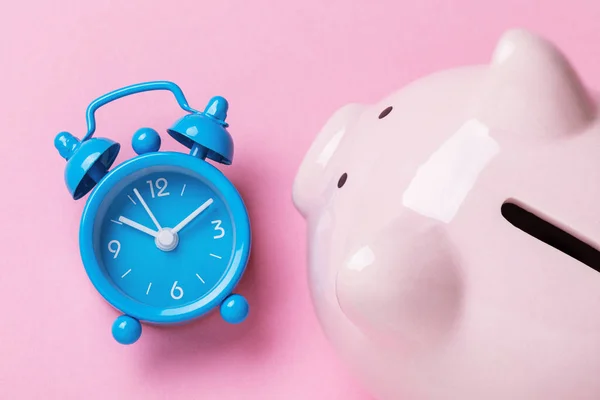 Alarm clock and piggy bank on a pink background, top view. Time